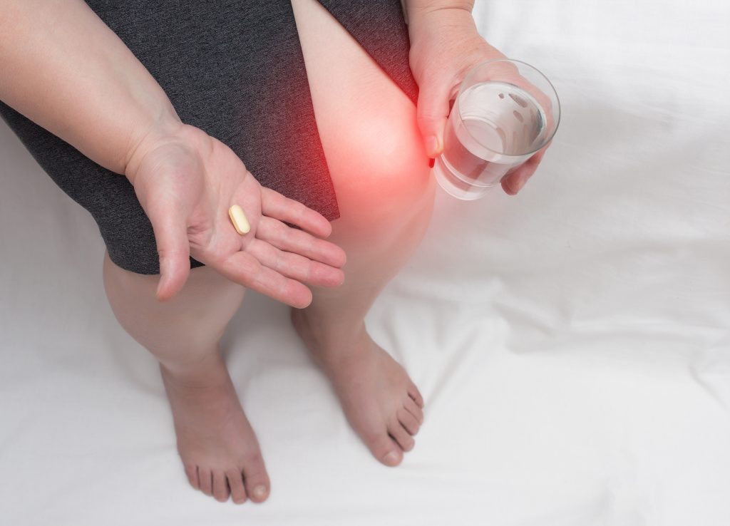 Frau mit einem schmerzenden Knie hält eine Glucosamin- und Chondroitinsulfattablette und ein Glas Wasser in der Hand